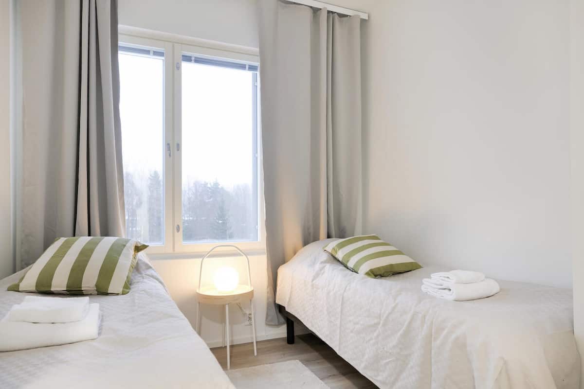 A room with two beds, each with a pillow and a folded towel. There is a small table with a lamp between the beds.