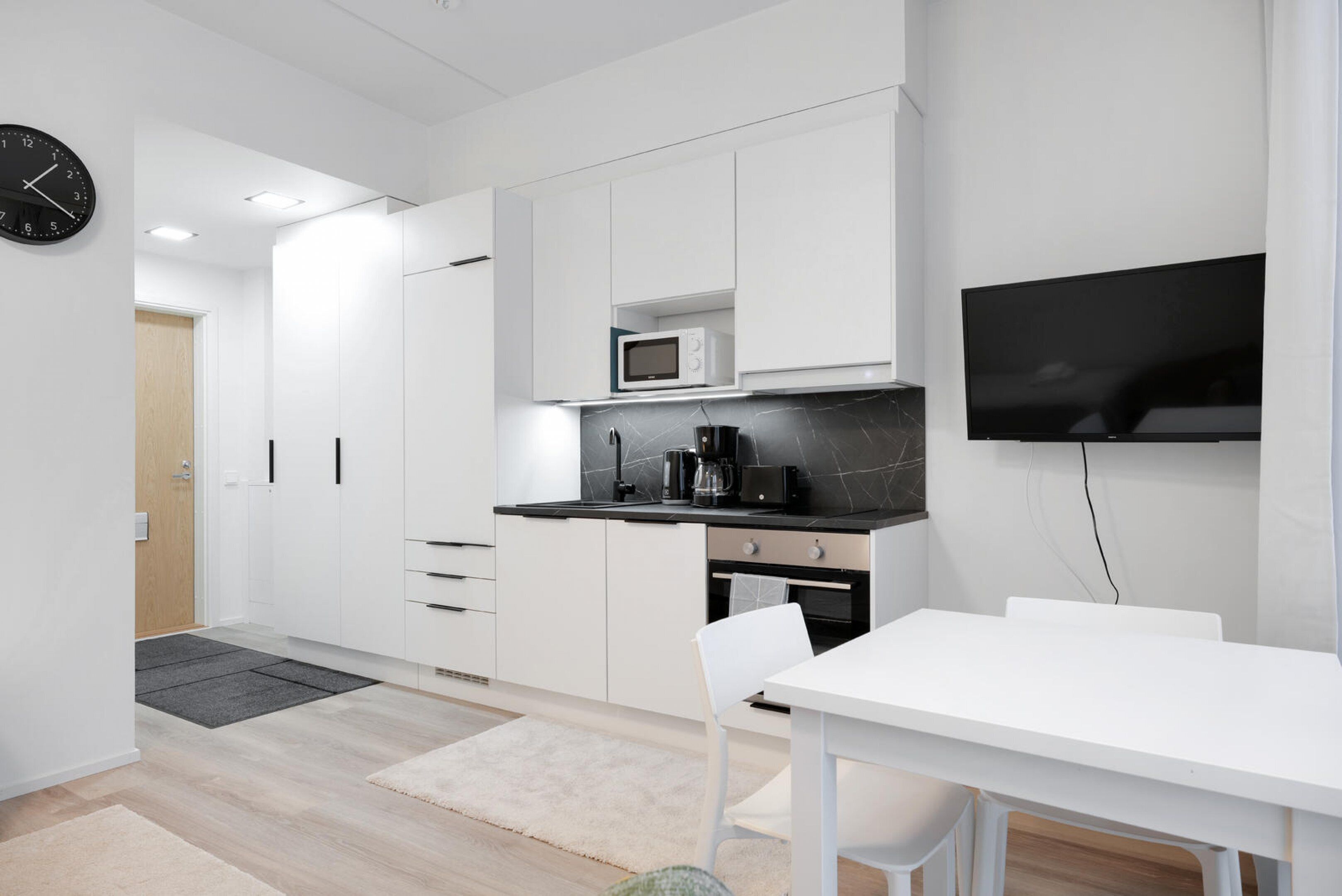Una cocina moderna con armarios blancos, microondas, cafetera y un televisor de pantalla plana montado en la pared.