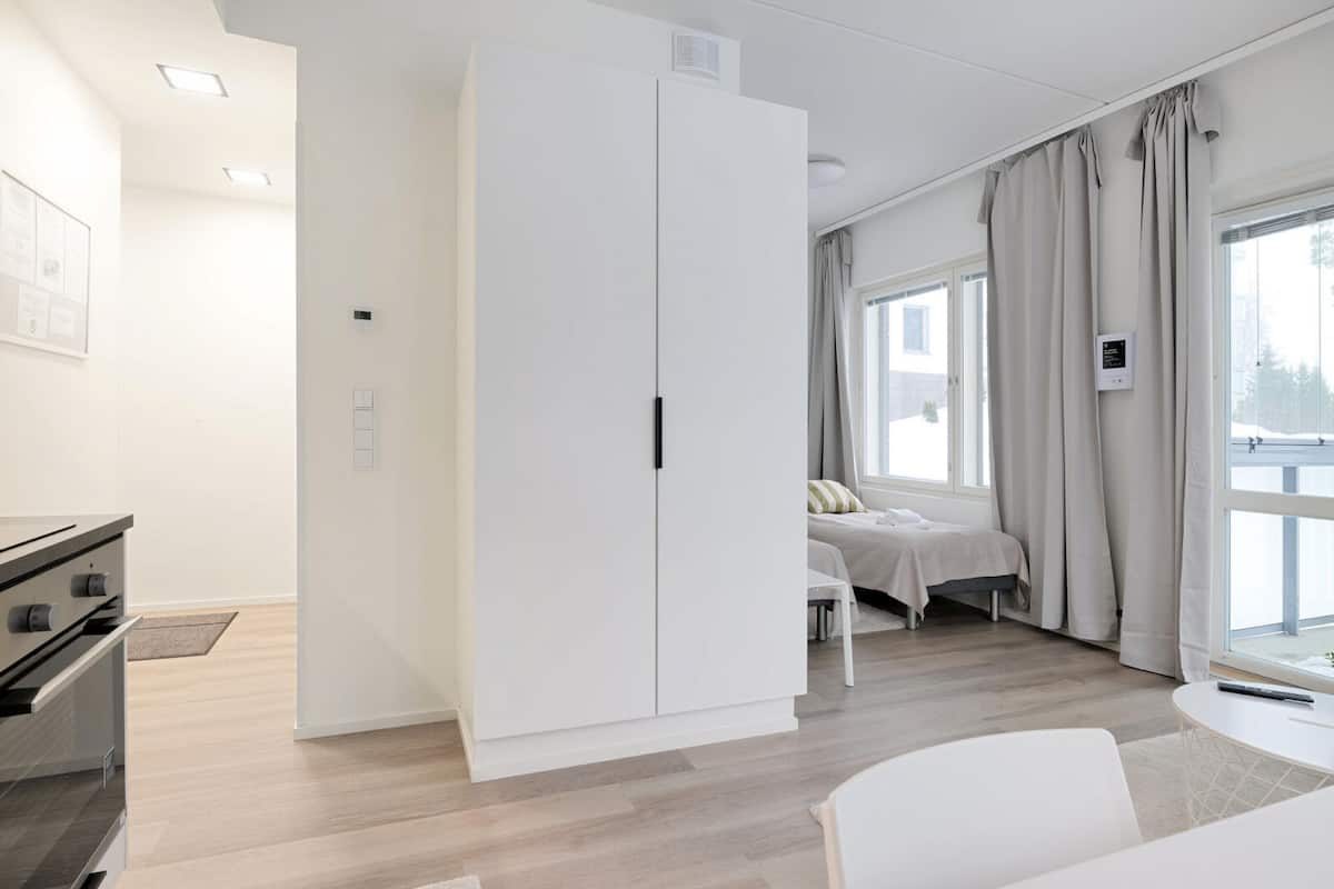 A modern hotel room with a large wardrobe, a bed with a white headboard, a small table, and a window with curtains.