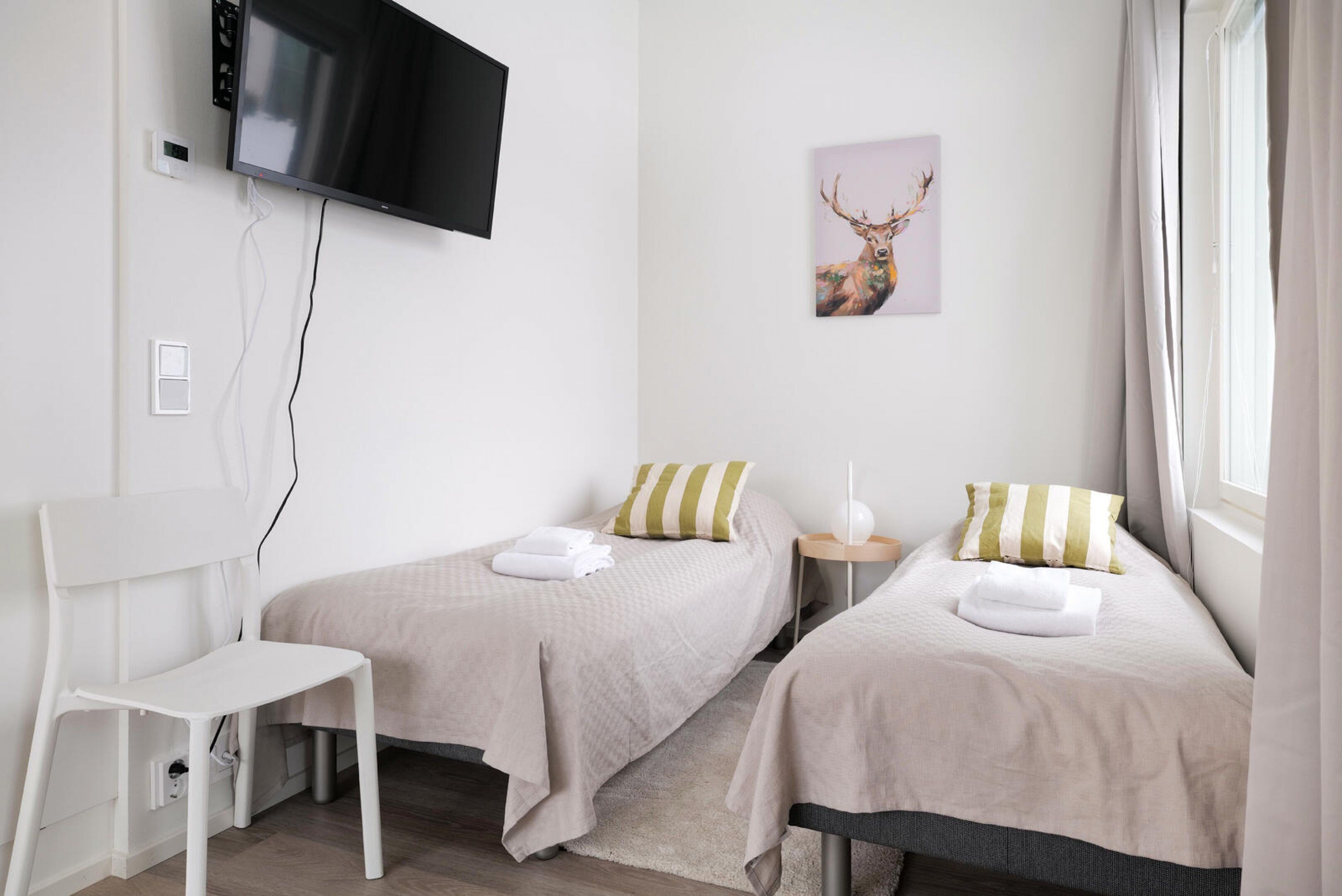 A hotel room with two beds, a TV mounted on the wall, a painting of a deer, and a white chair.