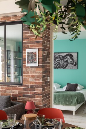 A modern room with a brick wall, a large mirror, a zebra artwork, a green bed, and a dining area with red chairs.