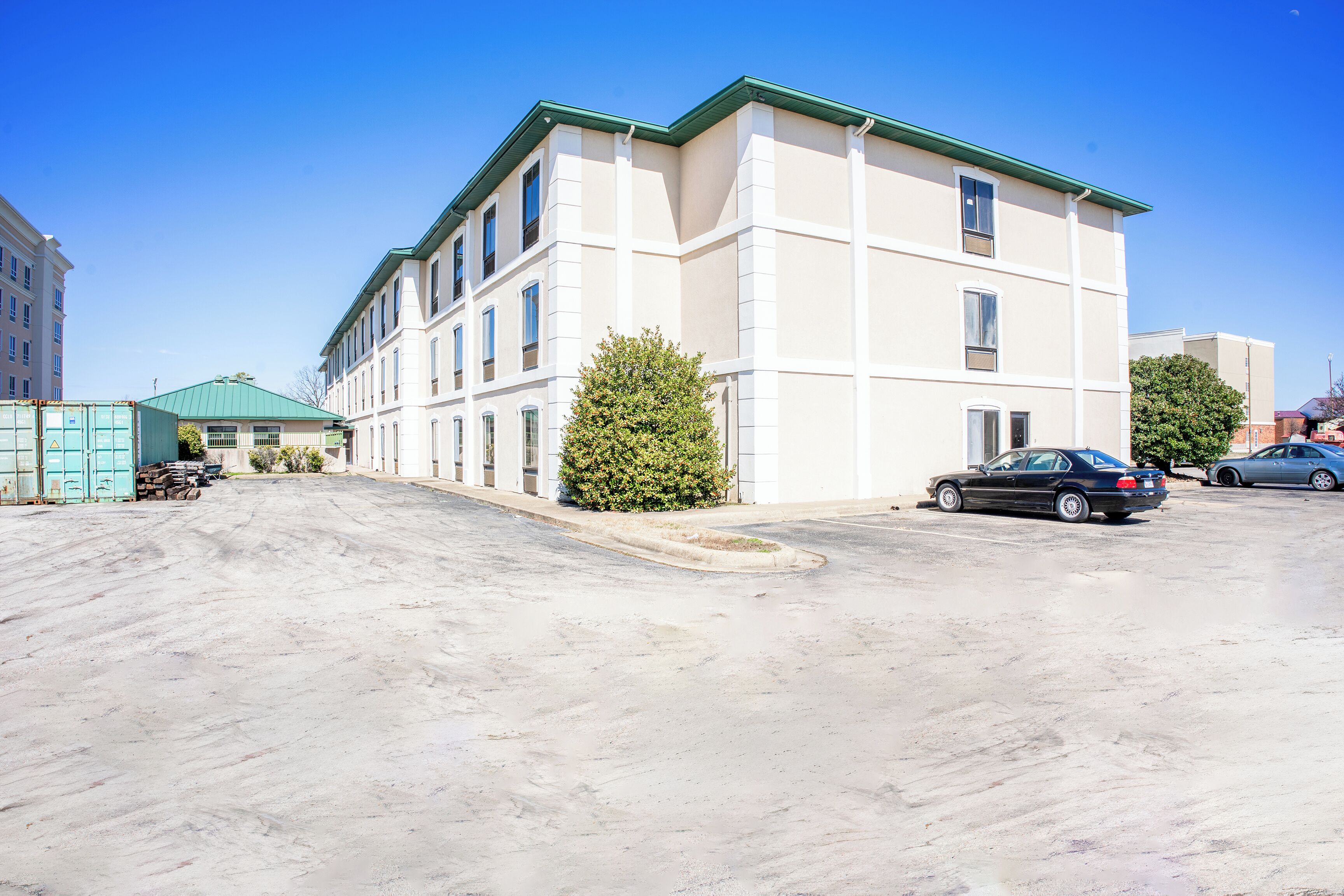 A multi-story building with a flat roof and a green trim. There are parked cars and a small tree in front.