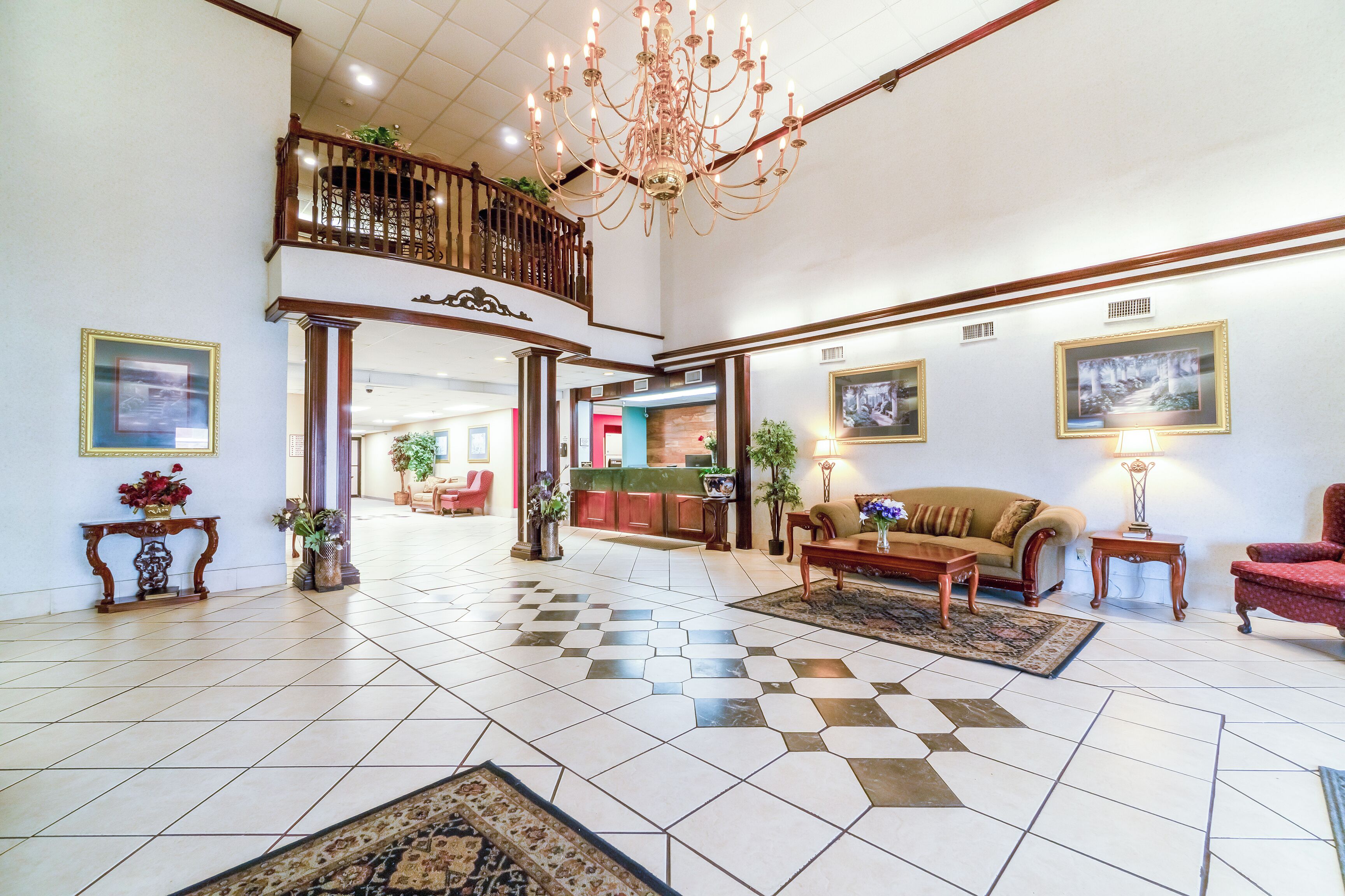 A spacious lobby with a checkered floor, a chandelier, a sofa, a red armchair, and framed pictures on the walls.