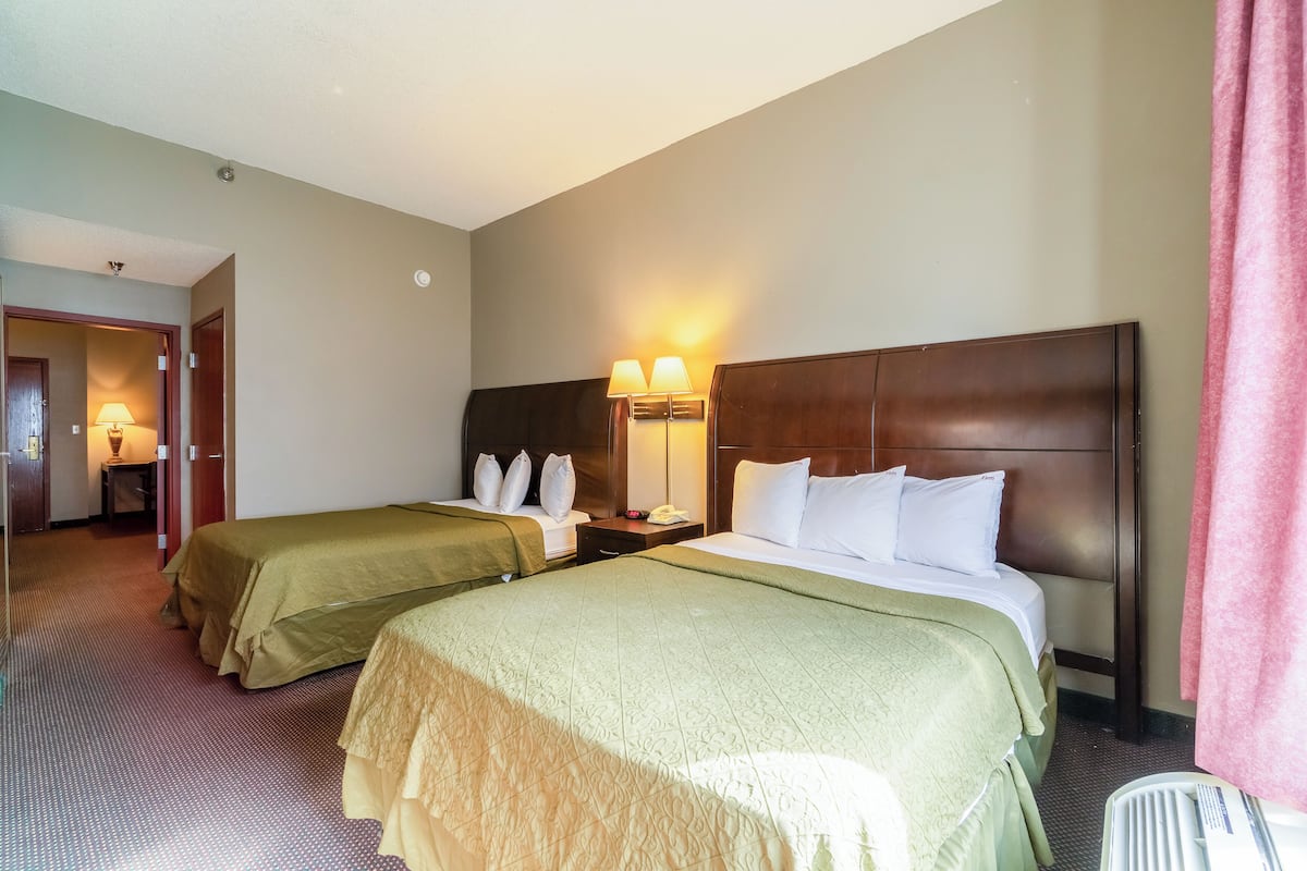 A hotel room with two beds, a wooden headboard, a nightstand, a lamp, and a bathroom area.