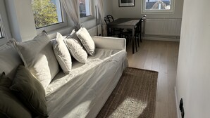 Una sala de estar con sofá, una mesa de comedor con sillas y amplias ventanas con cortinas.