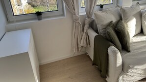 Habitación con sofá blanco, manta verde y vistas a un edificio y árboles a través de una ventana con cortinas atadas.