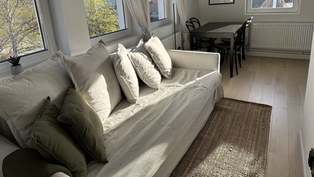 Una sala de estar moderna con un sofá, mesa de comedor, sillas y ventanas grandes con cortinas.