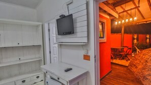 A compact room with a television mounted on the wall, a small white desk with a remote control, and a door leading to a dining area with a table and chairs. - La Gloria House (Ilhabela)