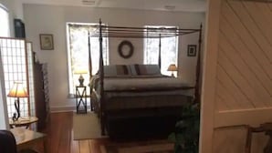 A bedroom with a four-poster bed, wooden flooring, a small table with a lamp, and a window with a view of trees. - 1831 House in Historic Old Towne Petersburg, VA (Petersburg)