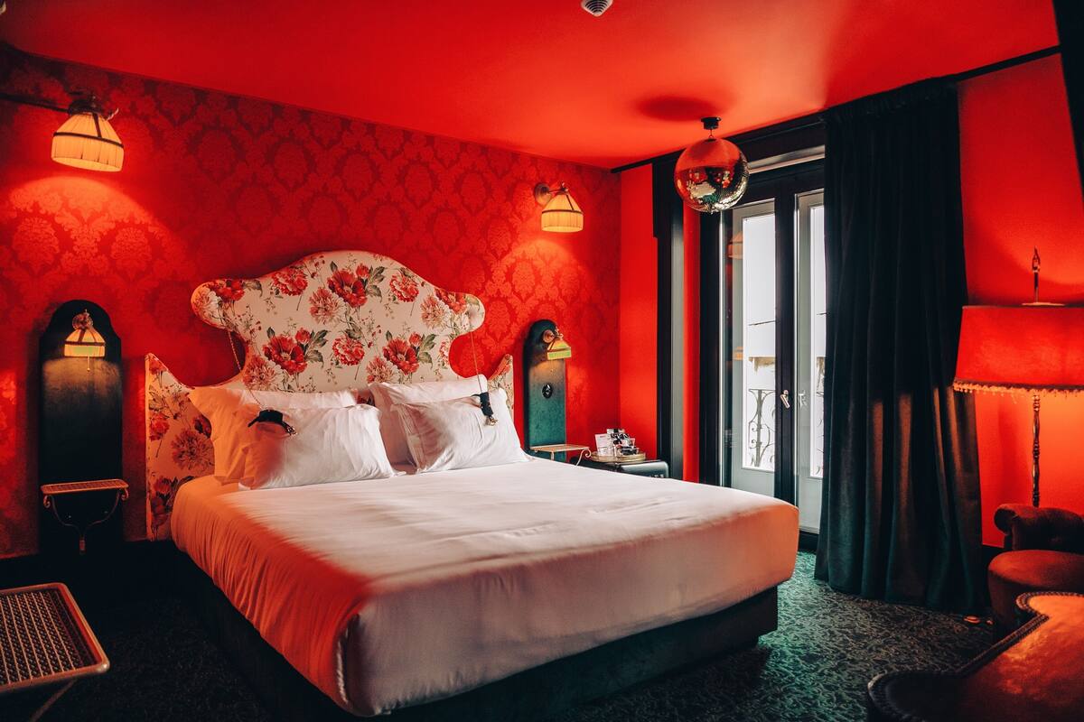 Habitación de hotel con una cama grande, cabecera floral, paredes rojas y dos lámparas fijadas a la pared.