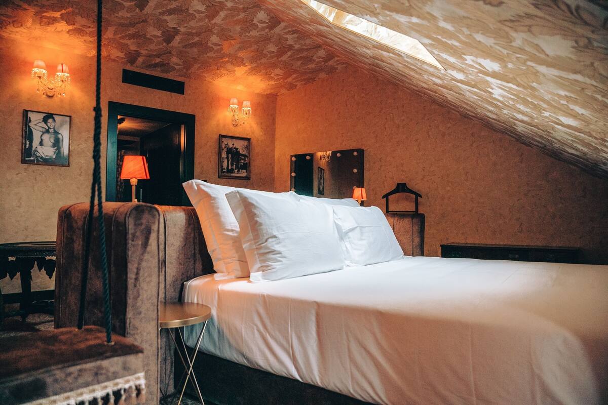 Una habitación acogedora y bien iluminada, con una cama, una silla y una mesa. Hay apliques y una araña que proporcionan luz.