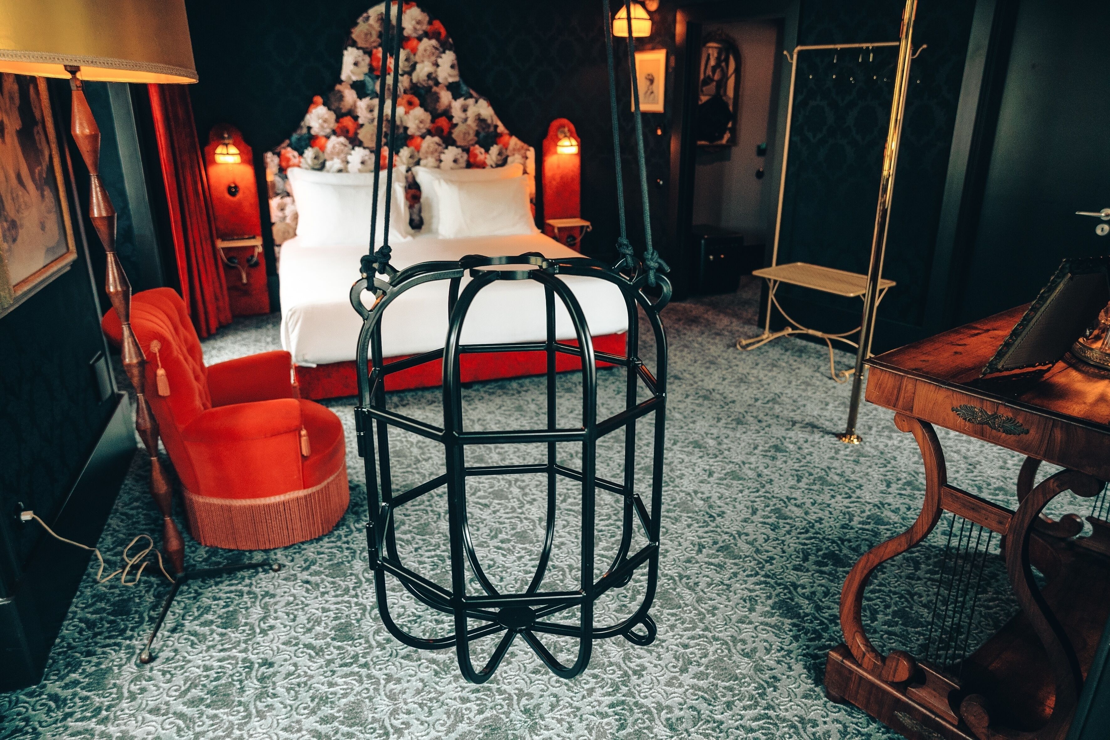 A hotel room with a bed, a red chair, a hanging chair, a music stand, and a lamp.