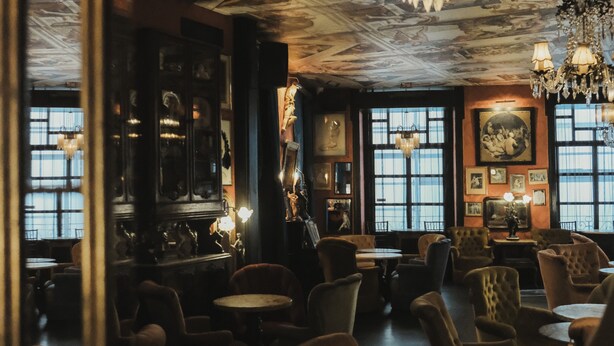 A dimly lit room with vintage decor, featuring multiple seating areas, a bar, and large windows. - Pensão Amor Madam’s Lodge (Lisbon)