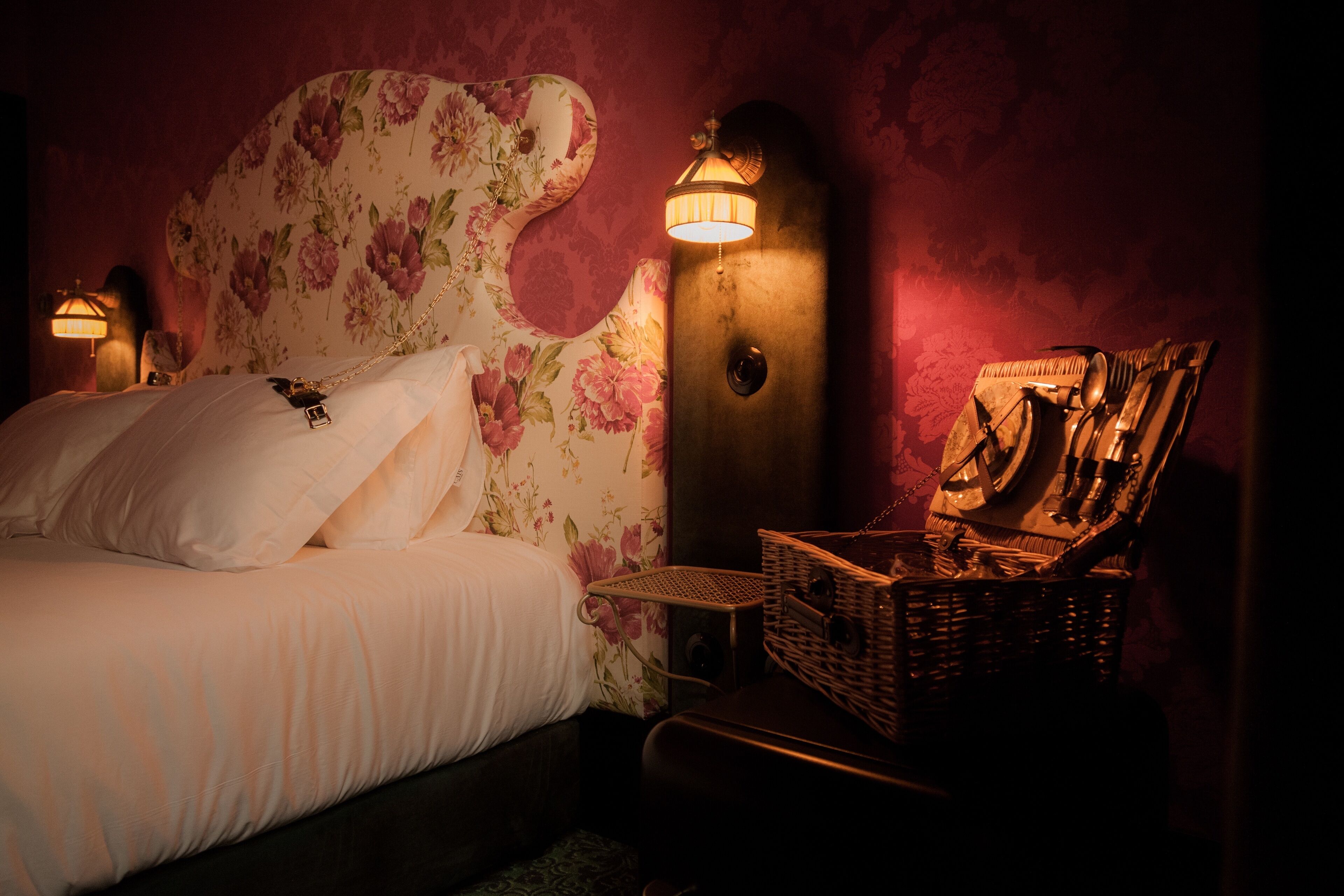 Ein Schlafzimmer mit einem blumigen Kopfteil, einer Nachttischlampe, einem Korbwäsches und einem gerahmten Bild an der Wand.