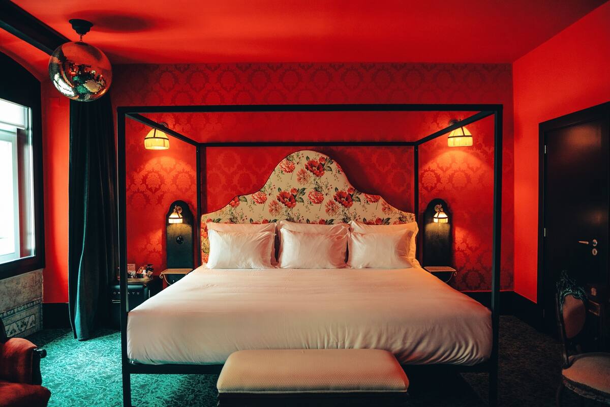 Una cama con dosel, cabecera con motivos florales, dos lámparas de noche y un banco al pie de la cama.