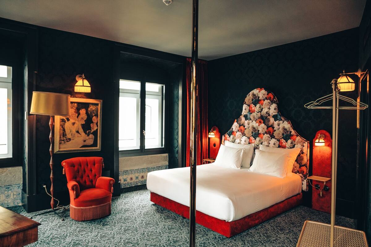 Habitación de hotel con una cama grande, un sillón rojo, una mesita de madera y una lámpara de pared.