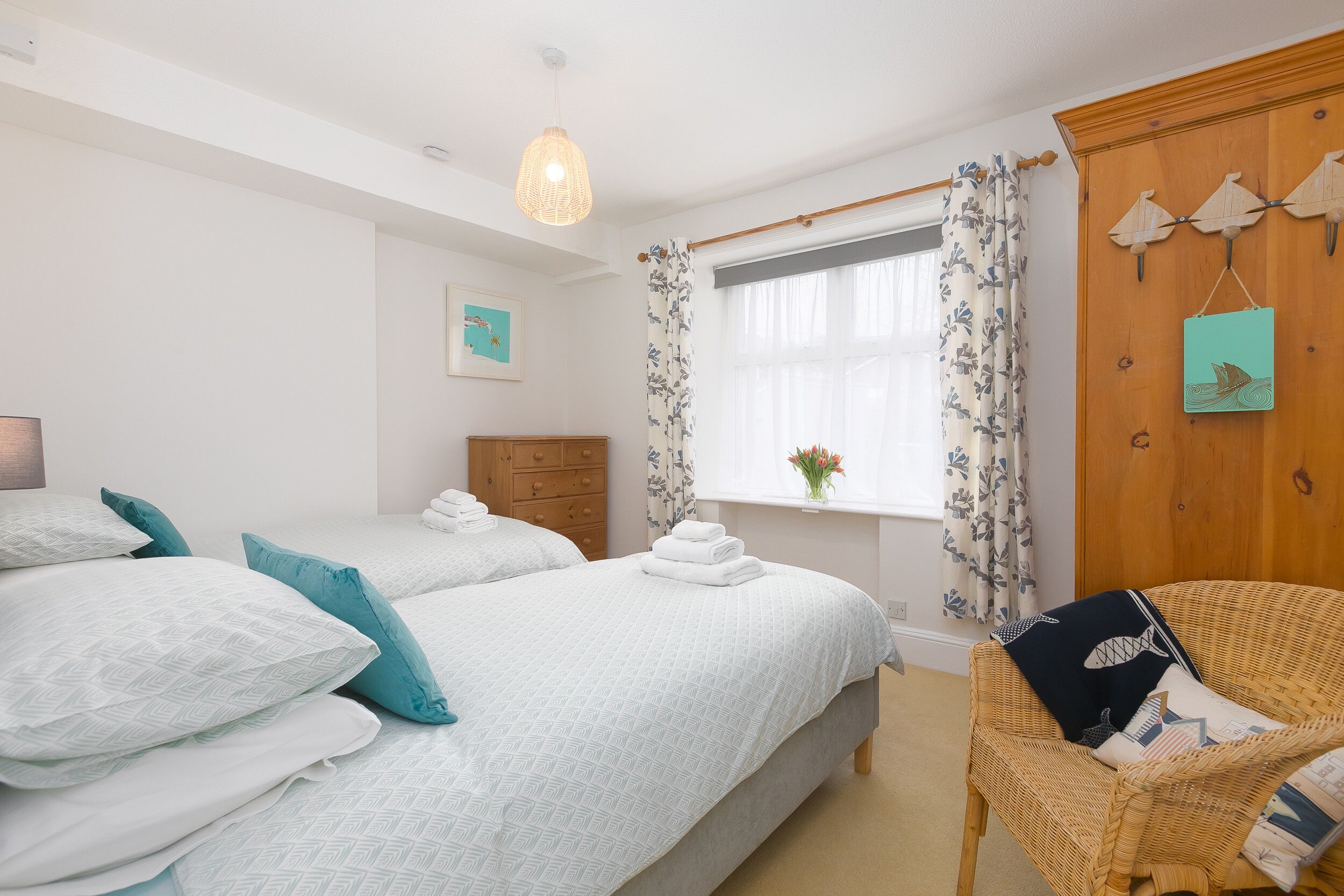 Une chambre avec deux lits, un fauteuil en osier, une fenêtre avec des rideaux et un élément décoratif fixé au mur.