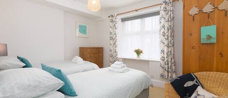 Une chambre avec deux lits, un fauteuil en osier, une fenêtre avec des rideaux et un élément décoratif fixé au mur.