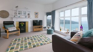Un salon avec une cheminée, un téléviseur, un canapé, une grande fenêtre donnant sur la mer et une horloge au mur.