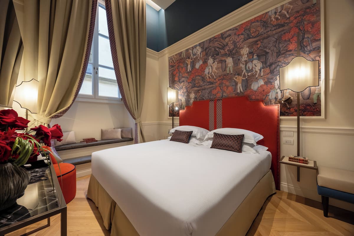 Une chambre d’hôtel avec un grand lit, une tête de lit rouge, une fresque murale, une fenêtre avec des rideaux et une table de chevet avec une lampe.