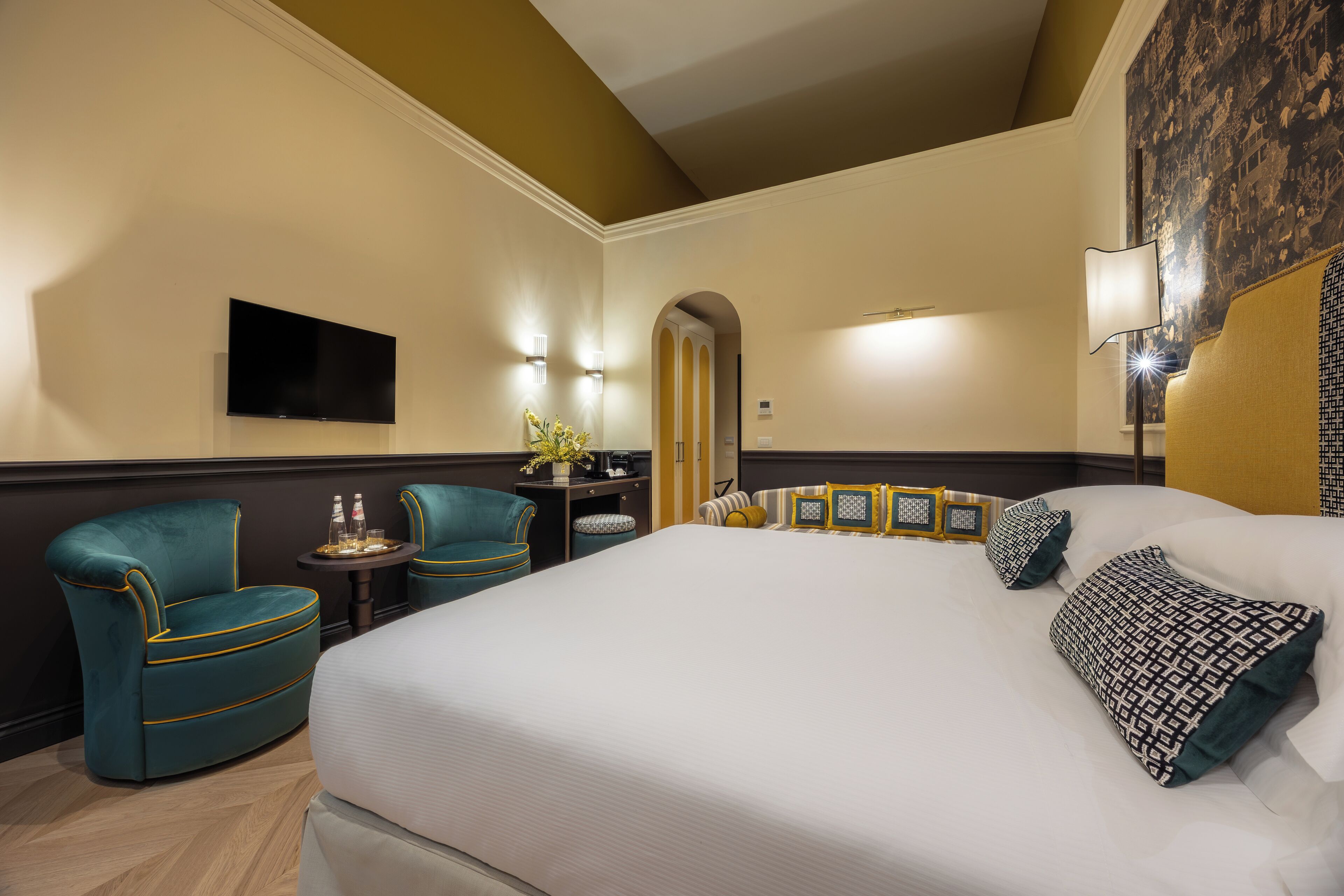 A hotel room with a large bed, two armchairs, a small table with a vase, and a flat-screen TV mounted on the wall.