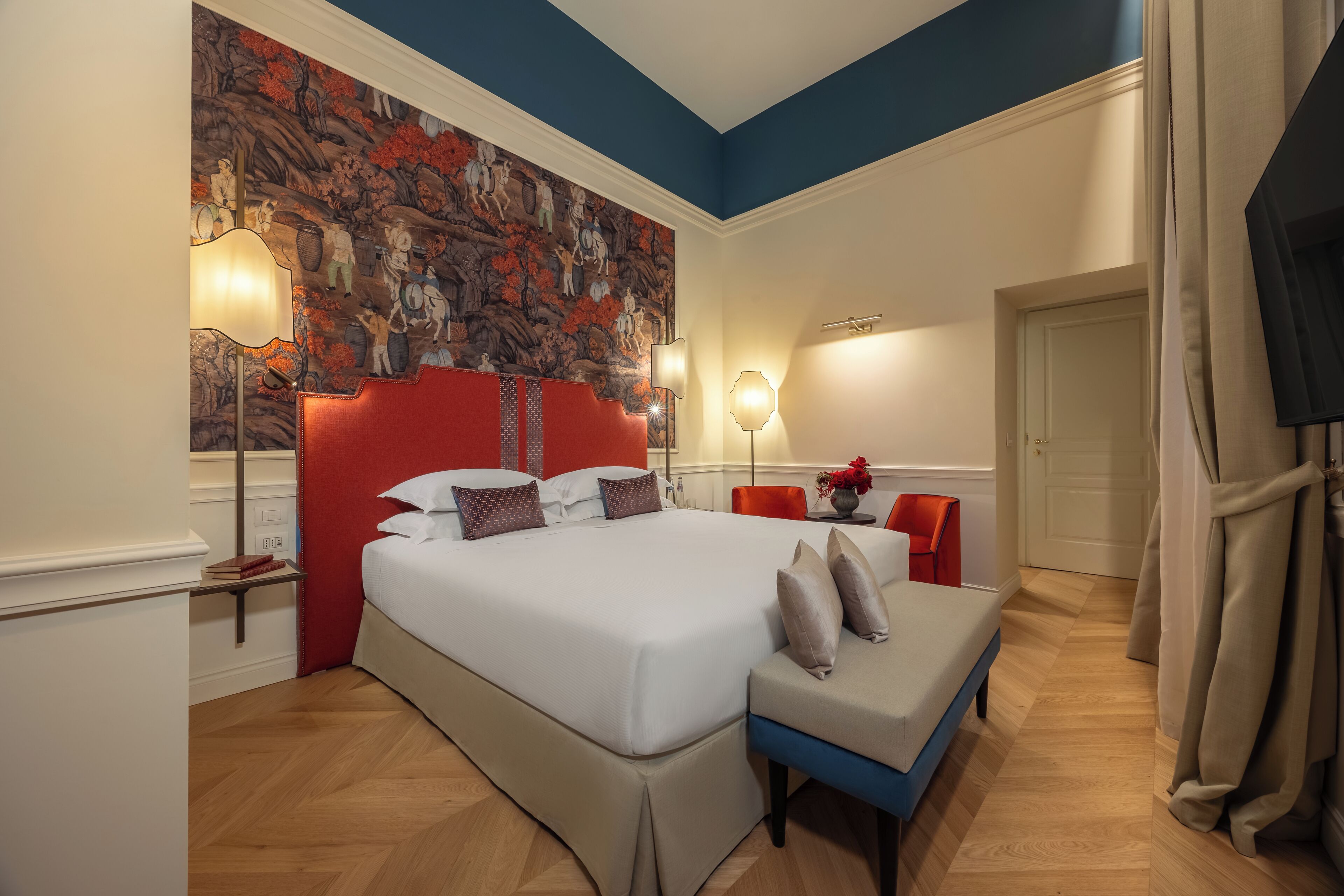 A hotel room with a large bed, a red headboard, a bench, a wall mural, and a door.