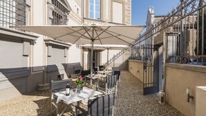 En uteservering med bord och stolar, ett stort parasoll och en byggnad med klassisk arkitektur.