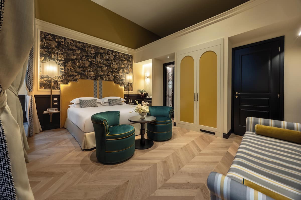 Une chambre d’hôtel avec un lit, deux fauteuils verts, un canapé à rayures, une petite table ronde avec un vase, et un grand miroir au-dessus du lit.