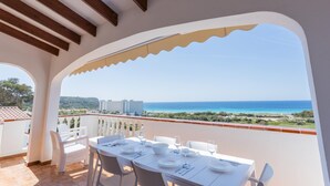 Un balcó amb taula preparada per dinar, amb vista al mar i edificis, i vegetació.