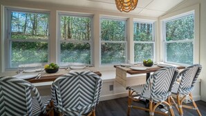 A dining area with a table set for four, surrounded by four chairs with patterned cushions, large windows with a view of trees, and a hanging light fixture. - Upscale Helen Cabin on 1 Wooded Acre (White County)