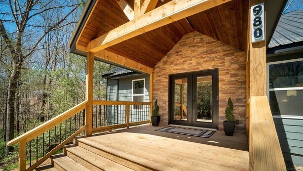 A wooden deck with a staircase leading to a house with a stone facade and a black door. - Upscale Helen Cabin on 1 Wooded Acre (White County)