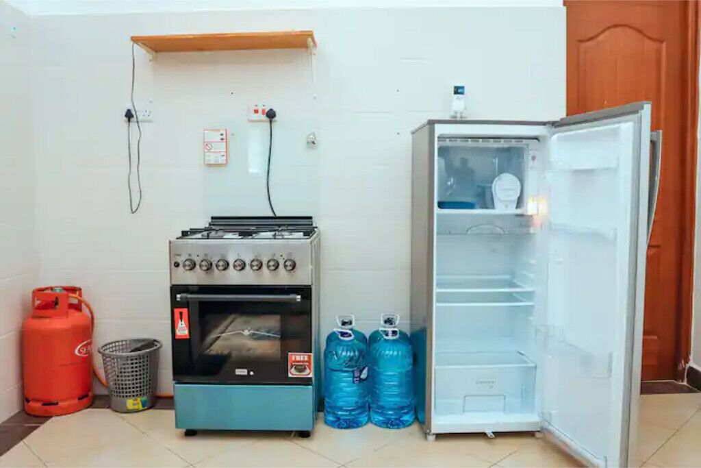 Eine kleine Küche mit Gasherd, Backofen und Kühlschrank. Zwei große Wasserkrüge und eine Gasflasche sind vorhanden.