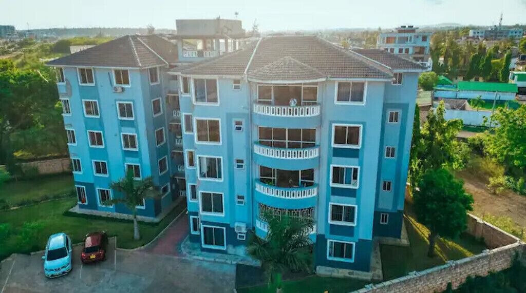 A multi-story residential building with balconies, a parking area, and surrounding greenery.