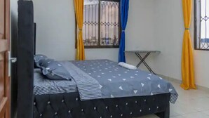 A bedroom with a bed, curtains, a window with bars, and a small table. - Divine apartments (Mombasa)