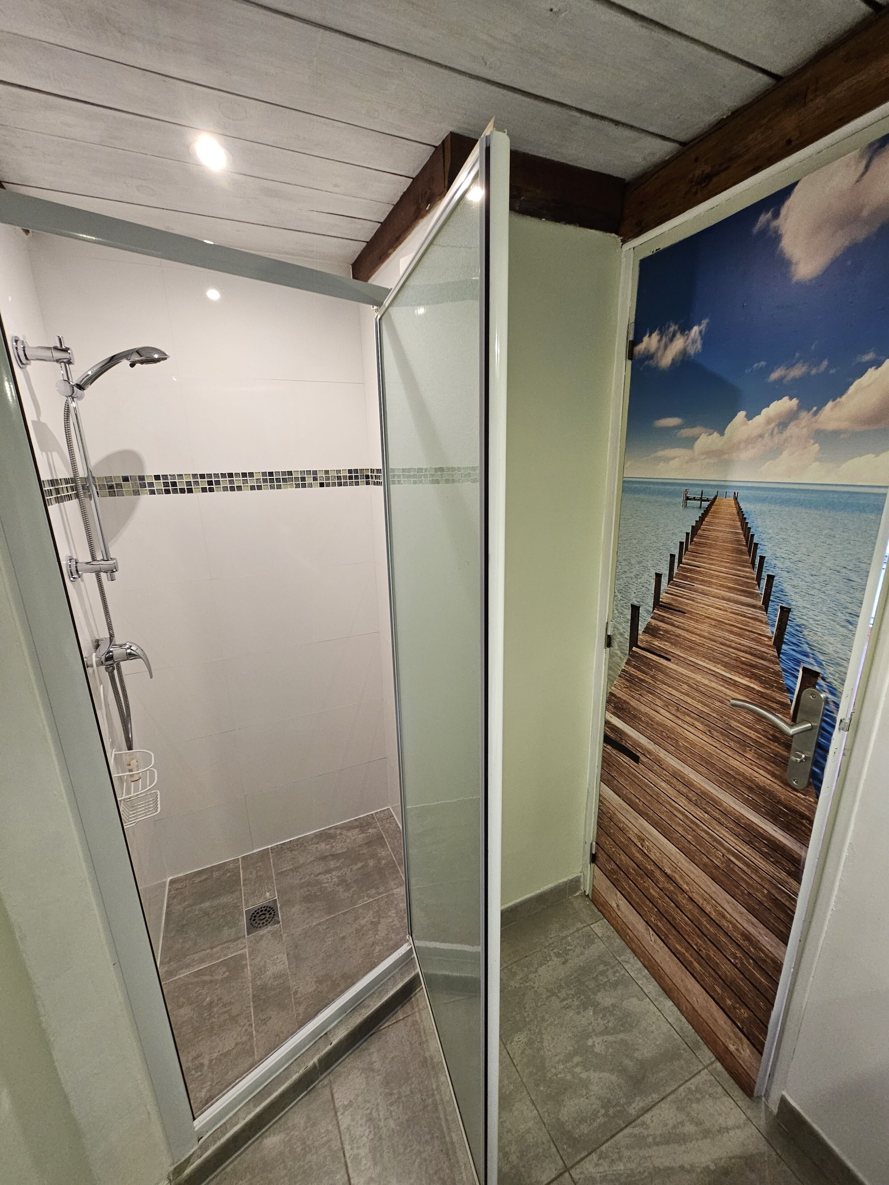 A bathroom with a glass shower enclosure, a tiled floor, and a wooden deck leading to a body of water.