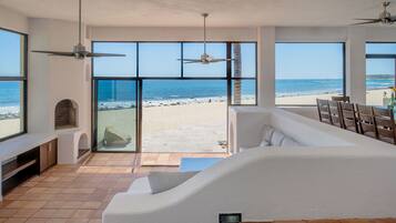 A spacious living area with large windows overlooking a beach, a ceiling fan, and a built-in fireplace.