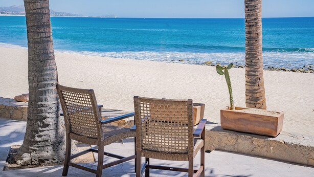 Two wicker chairs and a potted plant on a sandy beach with a clear blue ocean and palm trees in the background.