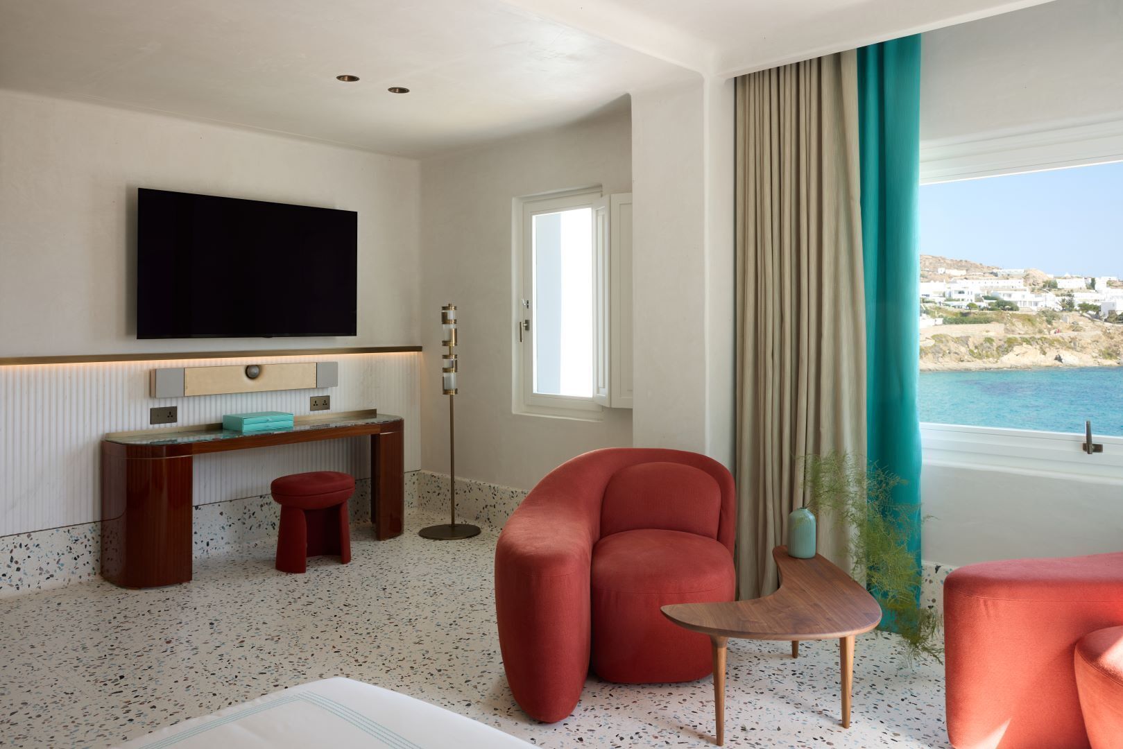 Habitación de hotel con una televisión grande, un escritorio, una silla roja, una mesita y vista al mar.