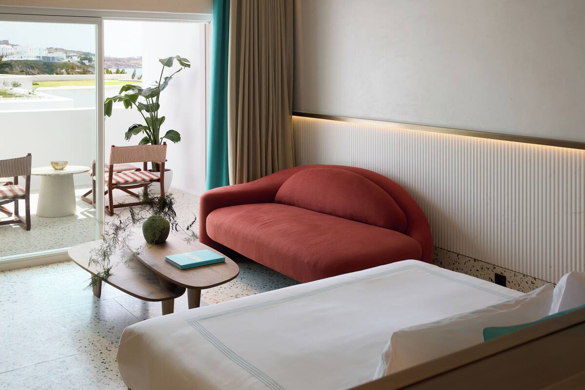 Habitación de hotel con cama, sofá rojo, una mesita con un libro, planta en maceta y vistas al exterior.
