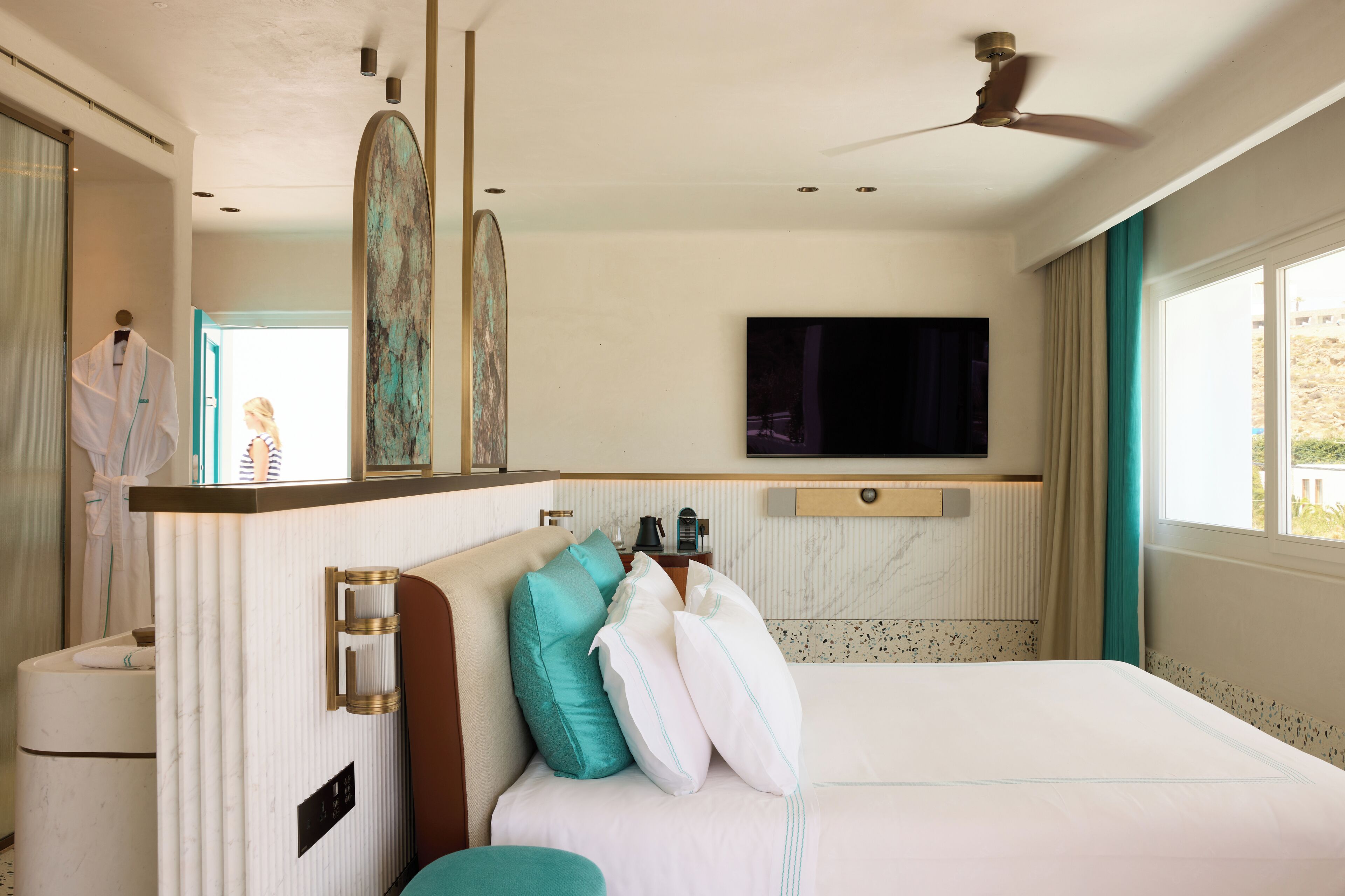 A hotel room with a large bed, a TV mounted on the wall, a ceiling fan, and a bathroom with a towel rack and sink.