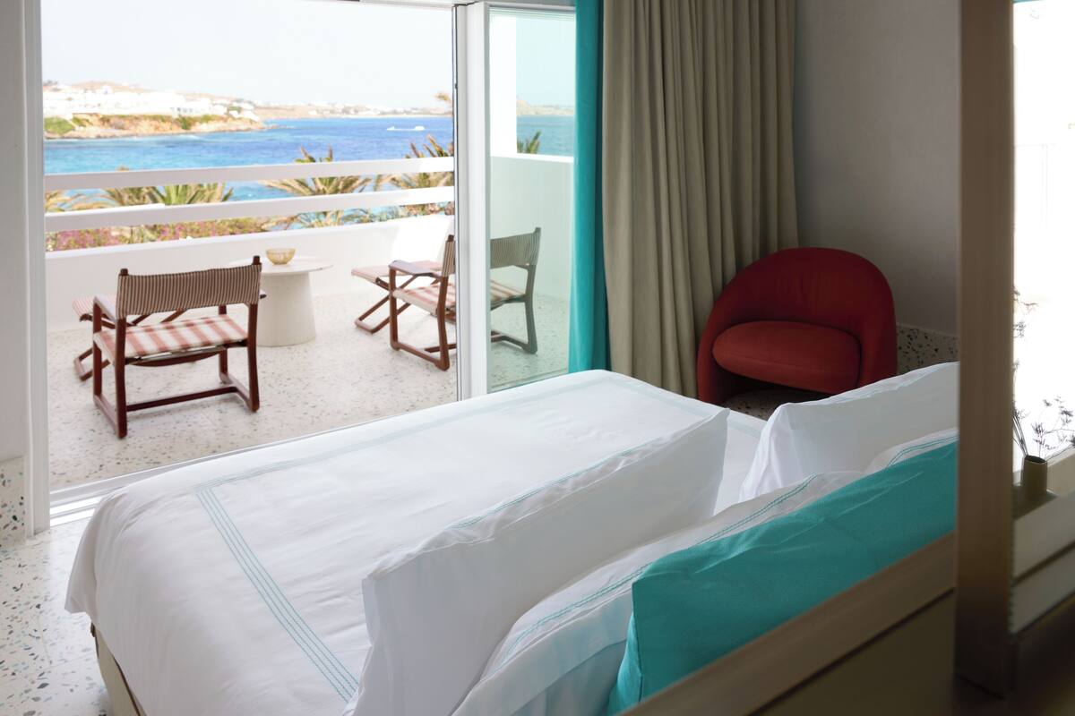 Habitación de hotel con una cama, una silla roja y vista al mar desde un balcón.