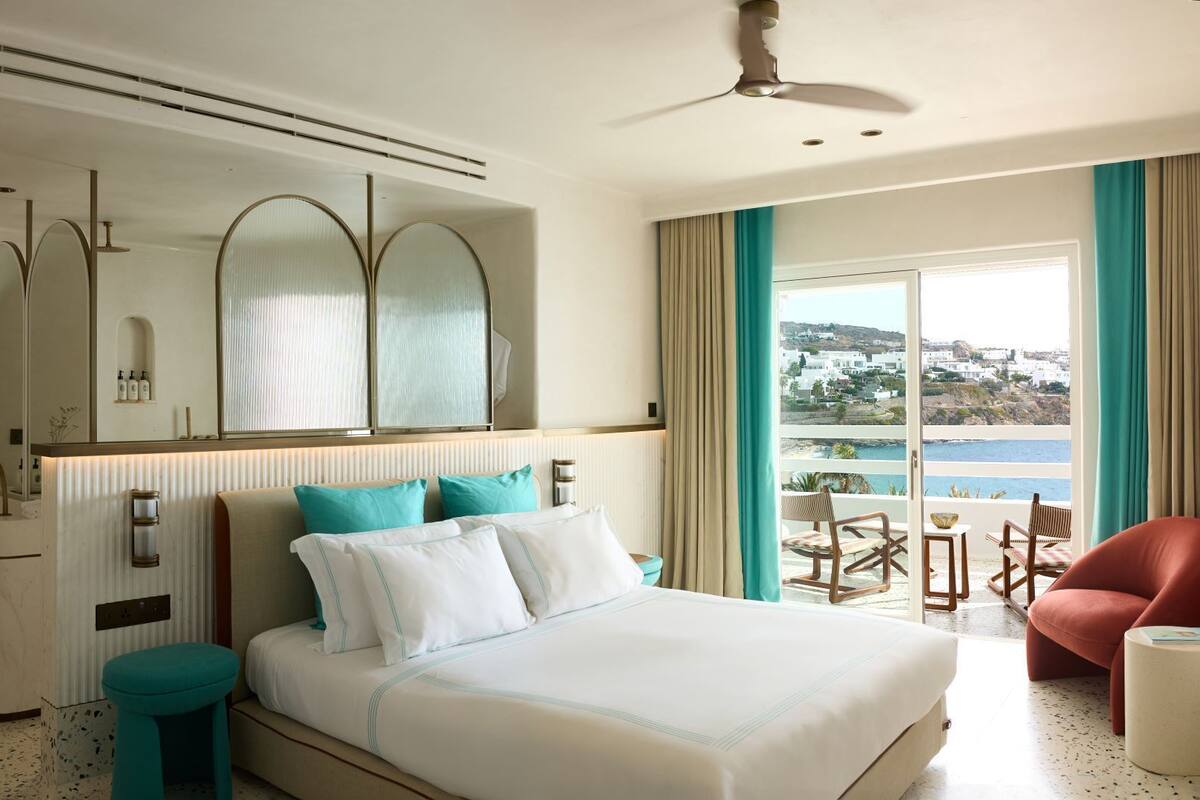 Habitación de hotel con cama, tocador con espejo, ventana con vista al mar y balcón con mobiliario exterior.
