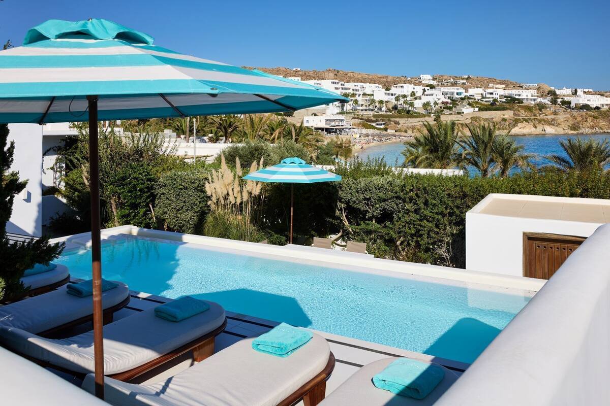 Zona de piscina con sillones reclinables y sombrillas, vista a un pueblo costero y cielo azul despejado.