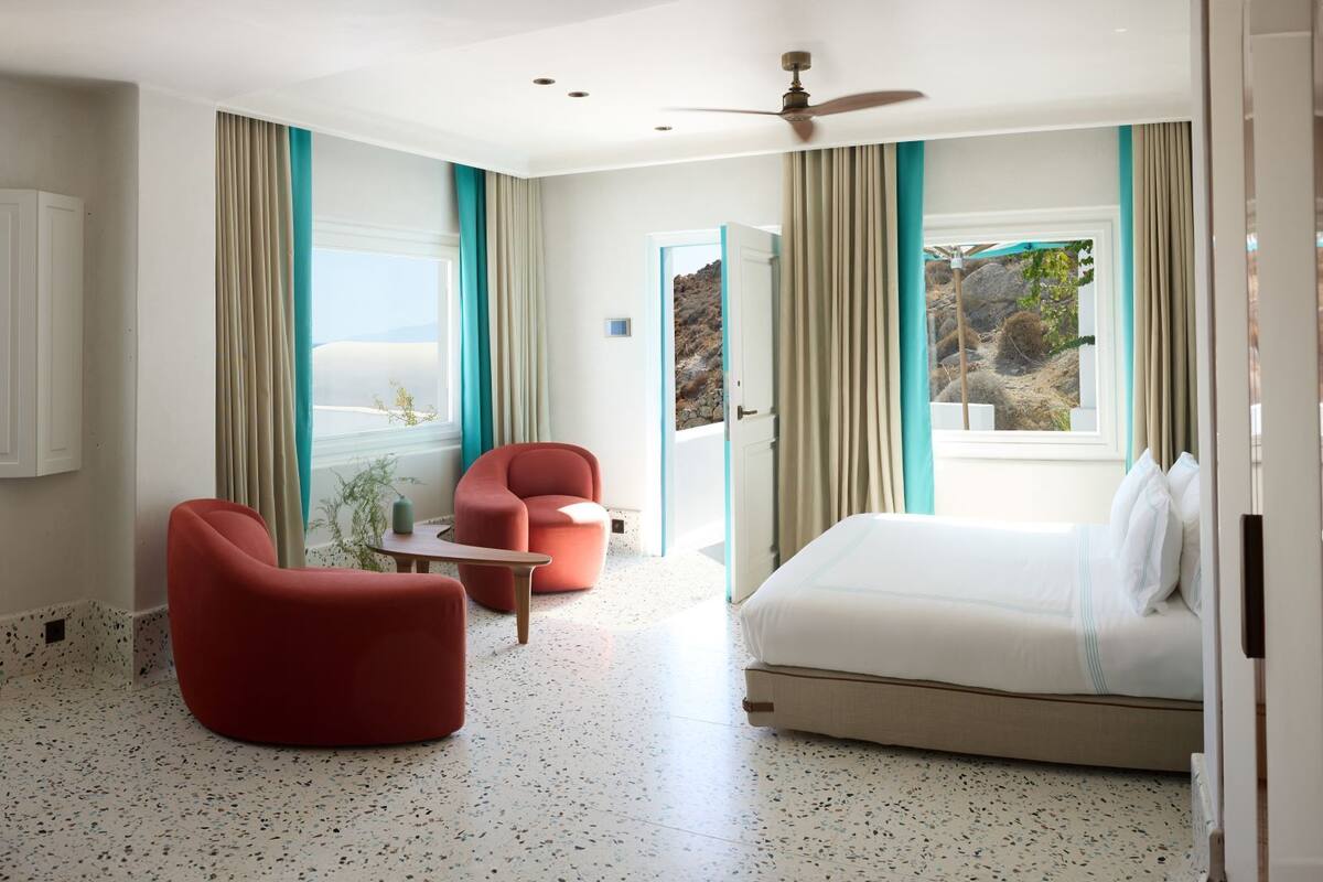 Habitación de hotel con una cama, dos sillas rojas, una mesita y un ventilador de techo, con vista al mar.