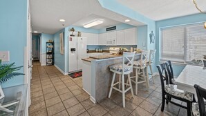 A compact kitchen with white cabinets, a central island with bar stools, and a dining area with a table and chairs. - Crescent Shores N 201-4BR Oceanfront Condo (North Myrtle Beach)