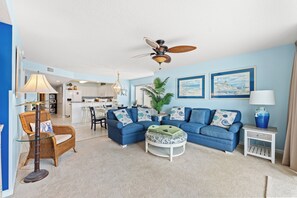 A living room with a blue sofa, a dining area, a ceiling fan, and two framed pictures on the wall. - Crescent Shores N 201-4BR Oceanfront Condo (North Myrtle Beach)