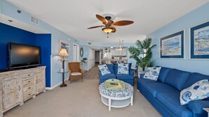A living room with a blue sofa, a television, a ceiling fan, and a dining area in the background. - Crescent Shores N 201-4BR Oceanfront Condo (North Myrtle Beach)