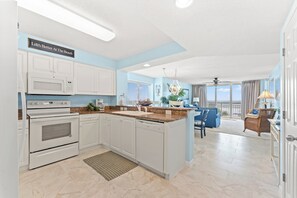 Eine moderne Küche mit weißen Schränken, einer eingebauten Mikrowelle und einem Backofen. Es gibt einen Essbereich mit einem blauen Stuhl und Blick auf den Strand durch große Fenster.