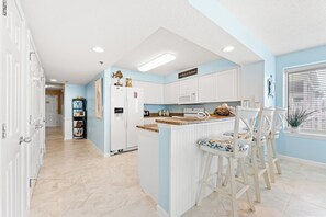A kitchen with white cabinets, a central island with bar stools, a microwave, and a refrigerator. - Crescent Shores N 201-4BR Oceanfront Condo (North Myrtle Beach)