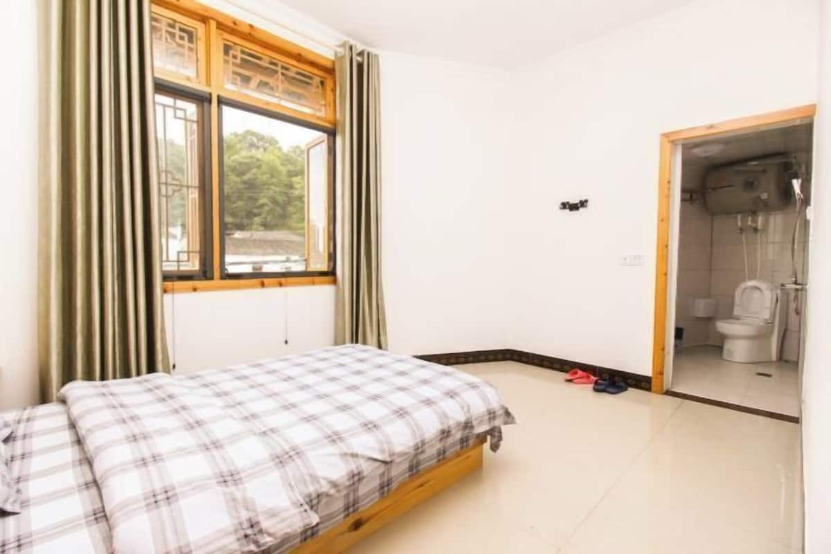 Quarto com cama, janela com cortinas, banheiro pequeno com vaso sanitário e chuveiro.